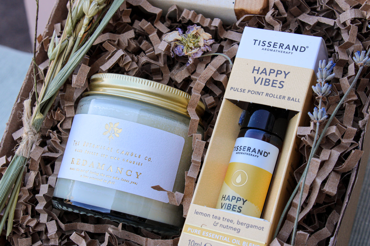 The Gardeners Delight Gift Box by Wild About Flowers includes a natural soy wax candle labeled "Redamancy" from The Botanical Candle Co. and a "Happy Vibes" pulse point roller ball from Tisserand Aromatherapy. It is beautifully decorated with dried flowers, lavender, and shredded paper for an extra special touch.