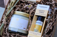 The Gardeners Delight Gift Box by Wild About Flowers includes a natural soy wax candle labeled "Redamancy" from The Botanical Candle Co. and a "Happy Vibes" pulse point roller ball from Tisserand Aromatherapy. It is beautifully decorated with dried flowers, lavender, and shredded paper for an extra special touch.
