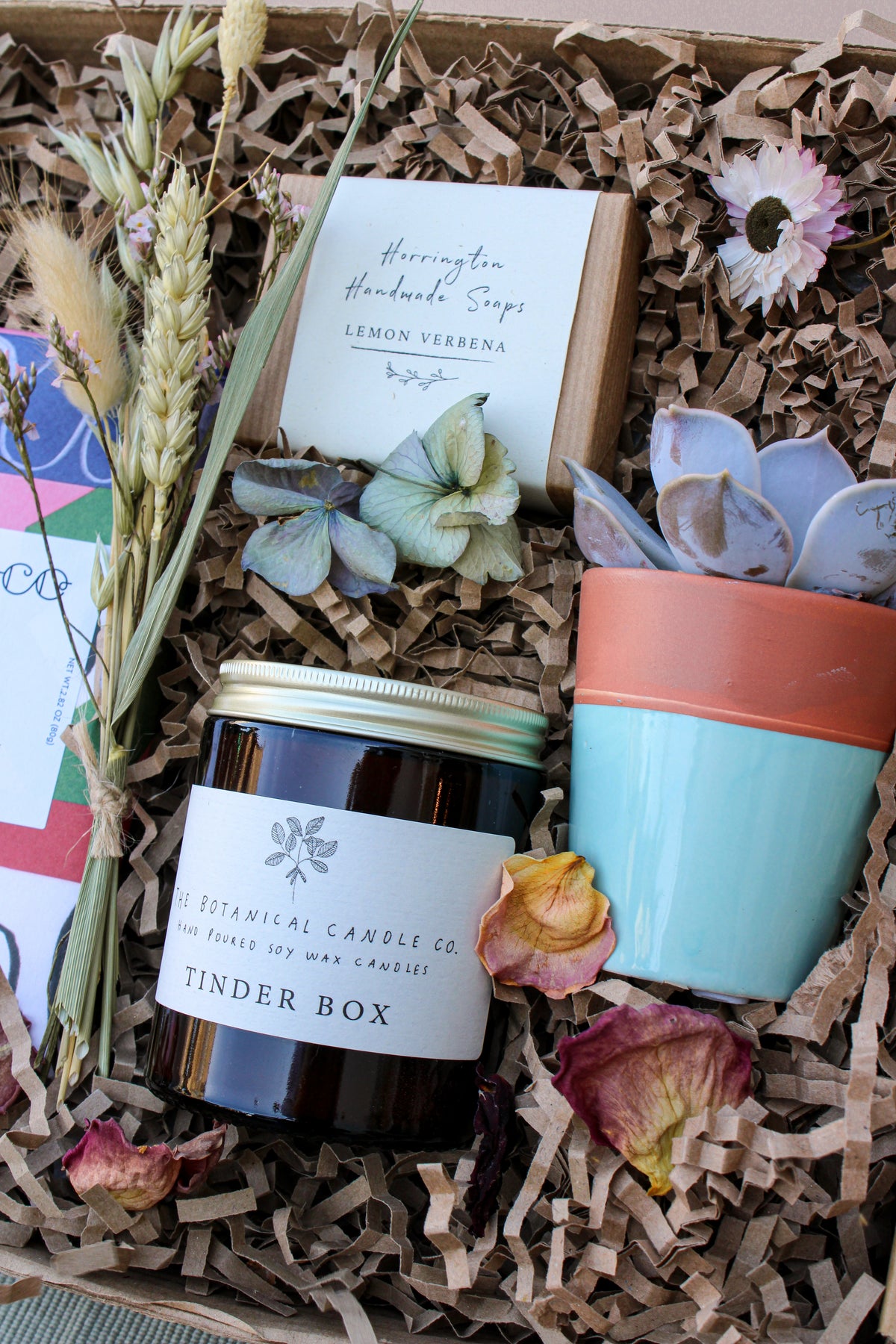 The Wild at Home Gift Box by Wild About Flowers is an eco-conscious set featuring a chic brown soy wax candle named "Tinder Box," a lemon verbena soap bar, a small pastel-leaf plant, dried flowers, and decorative shredded paper.