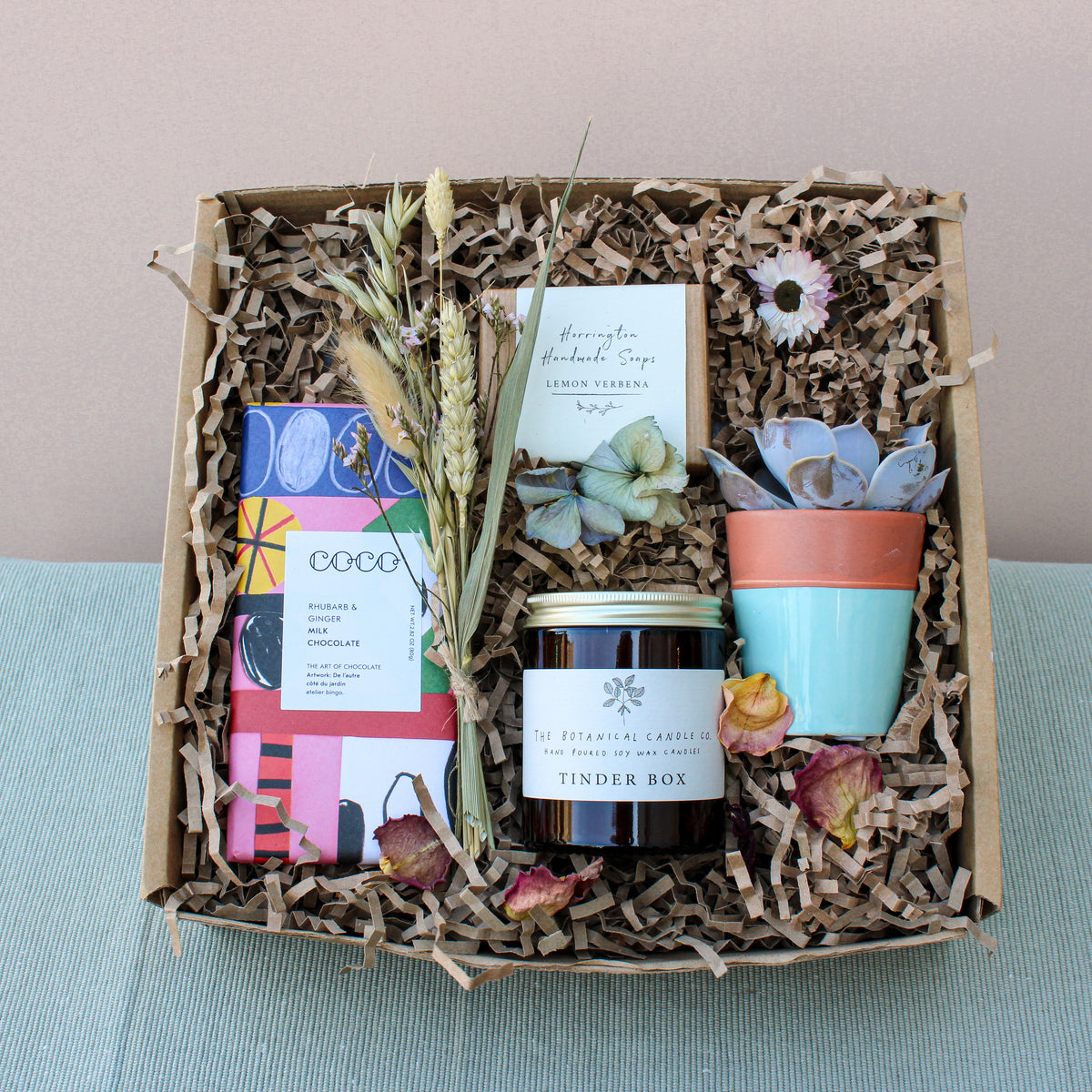 Explore the eco-friendly Wild at Home Gift Box by Wild About Flowers, showcasing a luxury chocolate bar, dried flowers, lemon verbena soap, and a succulent in a blue terracotta pot. Finished with a "Tinder Box" jar on shredded paper filler, it exudes thoughtful elegance.