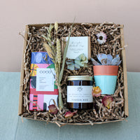Explore the eco-friendly Wild at Home Gift Box by Wild About Flowers, showcasing a luxury chocolate bar, dried flowers, lemon verbena soap, and a succulent in a blue terracotta pot. Finished with a "Tinder Box" jar on shredded paper filler, it exudes thoughtful elegance.