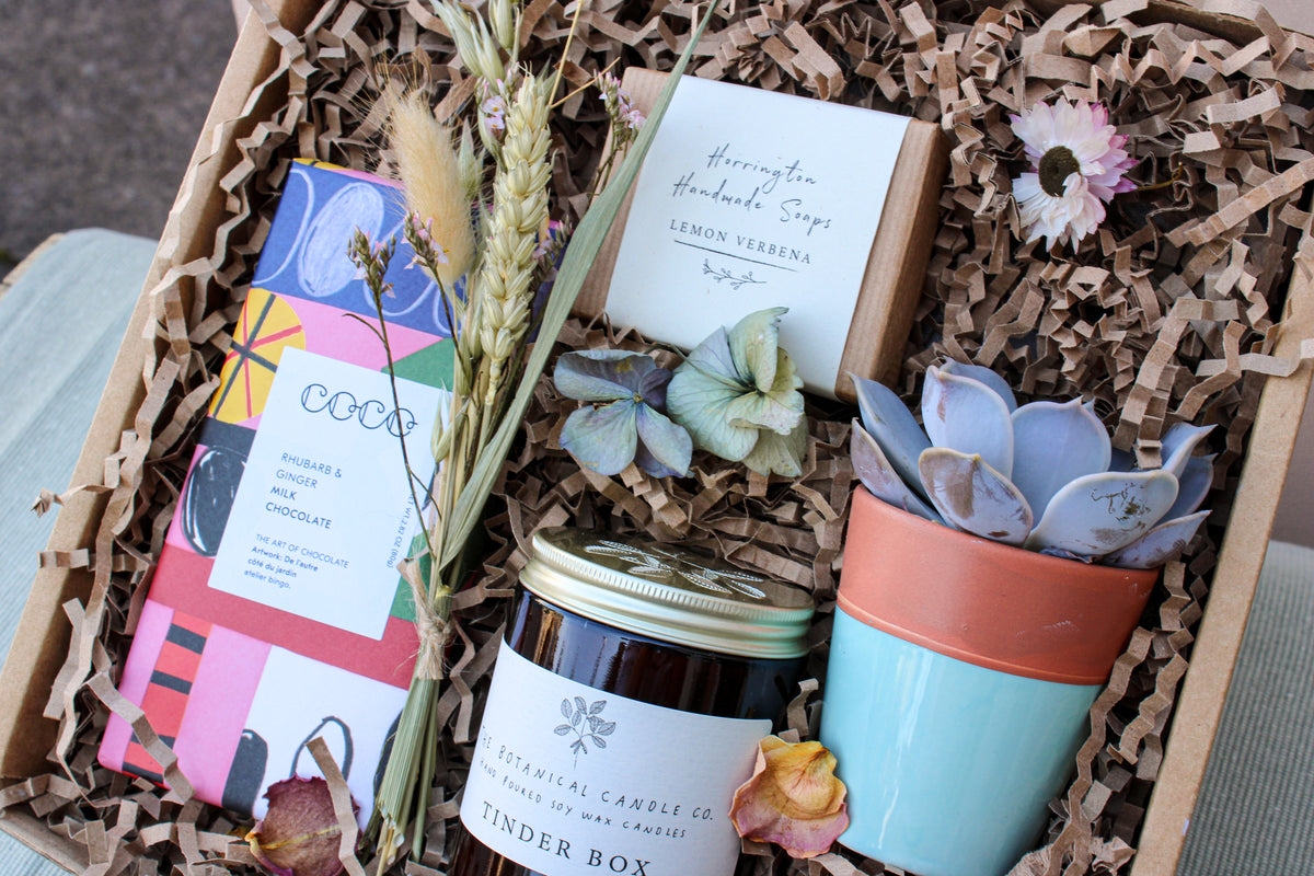 The Wild at Home Gift Box by Wild About Flowers includes a luxury chocolate bar, lemon verbena soap, a soy wax botanical candle, a potted succulent, dried flowers, and decorative paper shreds. Ideal for eco-conscious gifting.