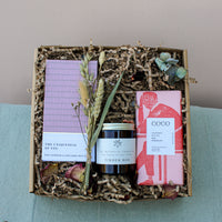 The Home Comforts Gift Box by Wild About Flowers includes a lavender striped notebook, a hand-poured soy wax candle named "Tinder Box," a red-packaged chocolate bar, and decorative dried flowers, all arranged on shredded paper lining to create an ideal cozy atmosphere.