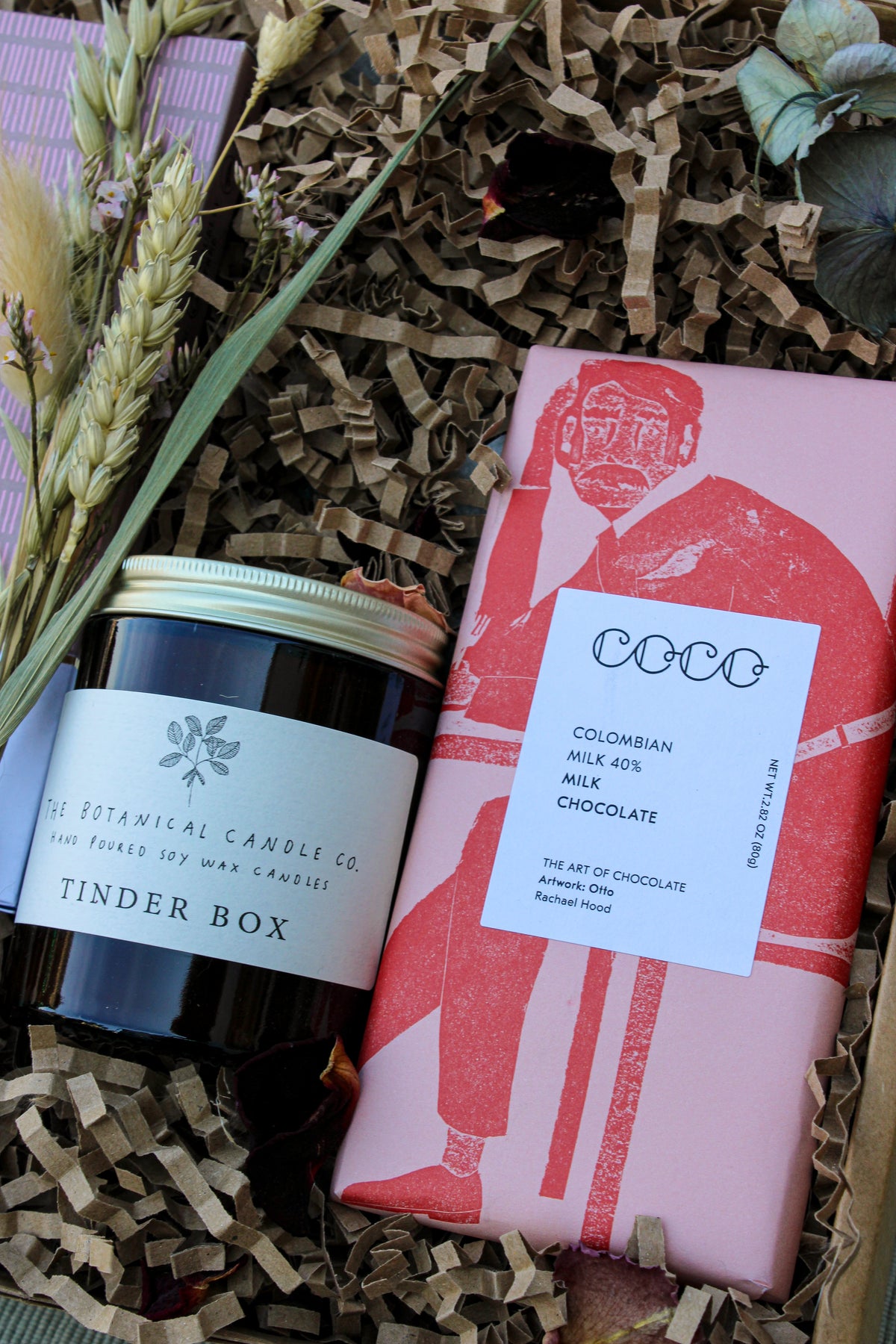 The Home comforts Gift Box by Wild About Flowers includes a hand-poured "Tinder Box" soy candle from The Botanical Candle Co. and a COCO Colombian milk chocolate bar, nestled in shredded paper with dried flowers.