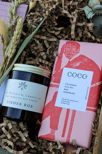 The Home comforts Gift Box by Wild About Flowers includes a hand-poured "Tinder Box" soy candle from The Botanical Candle Co. and a COCO Colombian milk chocolate bar, nestled in shredded paper with dried flowers.