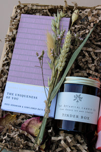 Introducing the Peace and Quiet Gift Box from Wild About Flowers: a tranquil assortment that includes "The Uniqueness of You" purple book, a jar candle labeled "Tinder Box" by The Botanical Candle Co., and a charming bouquet of dried flowers, all elegantly presented in eco-friendly shredded paper packaging. Ideal for creating an at-home spa experience.