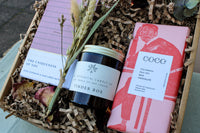 The Wild About Flowers Home Comforts Gift Box includes a pink book, "Tinder Box" hand-poured soy candle, and a pink chocolate bar, all nestled in shredded paper with dried flowers.