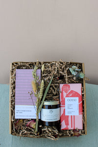 The Wild About Flowers Home Comforts Gift Box includes a book, bath oil bar, hand-poured soy candle, pink-wrapped chocolate, dried flowers, and brown shredded paper packing arranged on a light blue fabric.