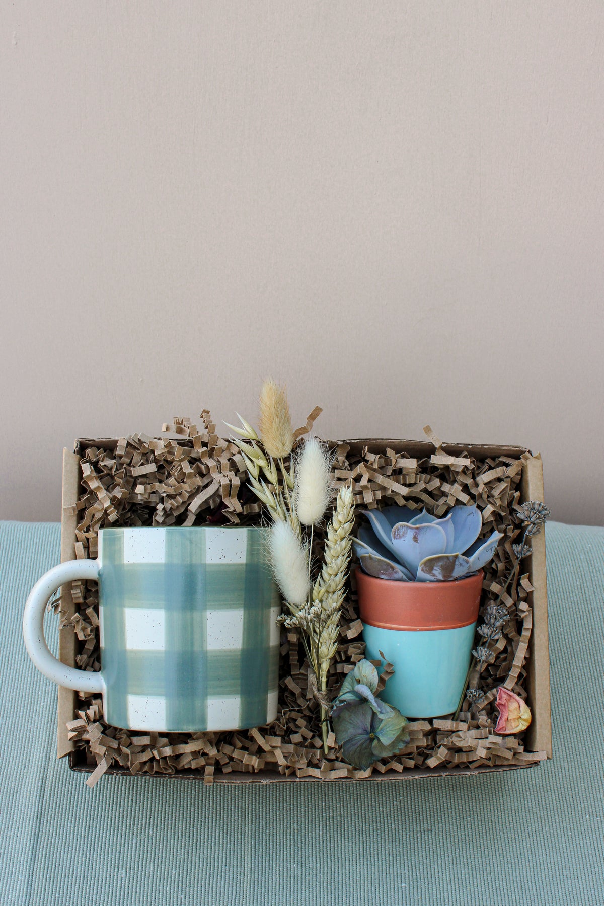 The Wild About Flowers New Housemates Gift Box includes a green and white checkered mug, a potted botanical plant, and dried flowers in eco-friendly packaging—a thoughtful housewarming gift.