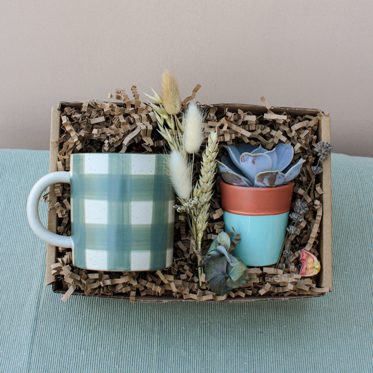 The Wild About Flowers New Housemates Gift Box features eco-friendly packaging with a green plaid mug, a potted botanical plant, and dried flowers—an ideal housewarming present set on light blue fabric.