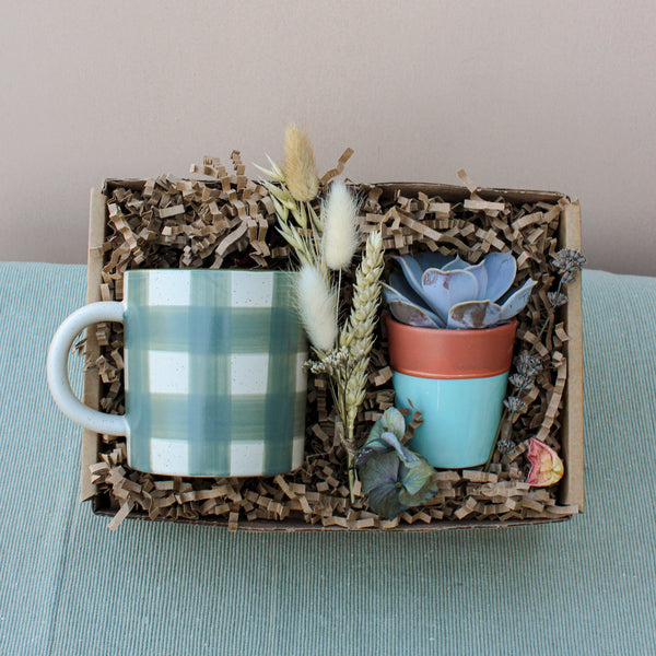 The Wild About Flowers New Housemates Gift Box features eco-friendly packaging with a green plaid mug, a potted botanical plant, and dried flowers—an ideal housewarming present set on light blue fabric.