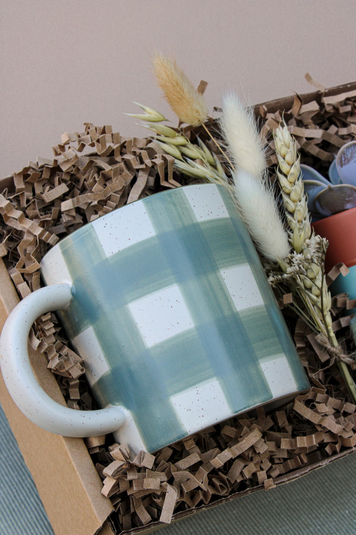 The New Housemates Gift box by Wild About Flowers features a white mug with a blue-green plaid pattern, eco-friendly packaging, dried grasses, and a botanical plant—an ideal housewarming gift.