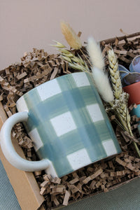 The New Housemates Gift box by Wild About Flowers features a white mug with a blue-green plaid pattern, eco-friendly packaging, dried grasses, and a botanical plant—an ideal housewarming gift.