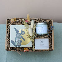 The Wild About Flowers New Mama Gift Box features an abstract black and gold card, dried flower bouquet, "Quiescent" candle, hypo-allergenic WS+Co MAMA + BABY soap, and decorative paper shreds for a relaxing gift experience.