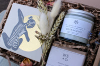 Wild About Flowers' New Mama Gift Box includes a card with an illustrated person on a yellow circle, a "Quiescent" candle for relaxation, dried flowers, hypo-allergenic soap, and WS+Co Mama & Baby product on brown crinkle paper.