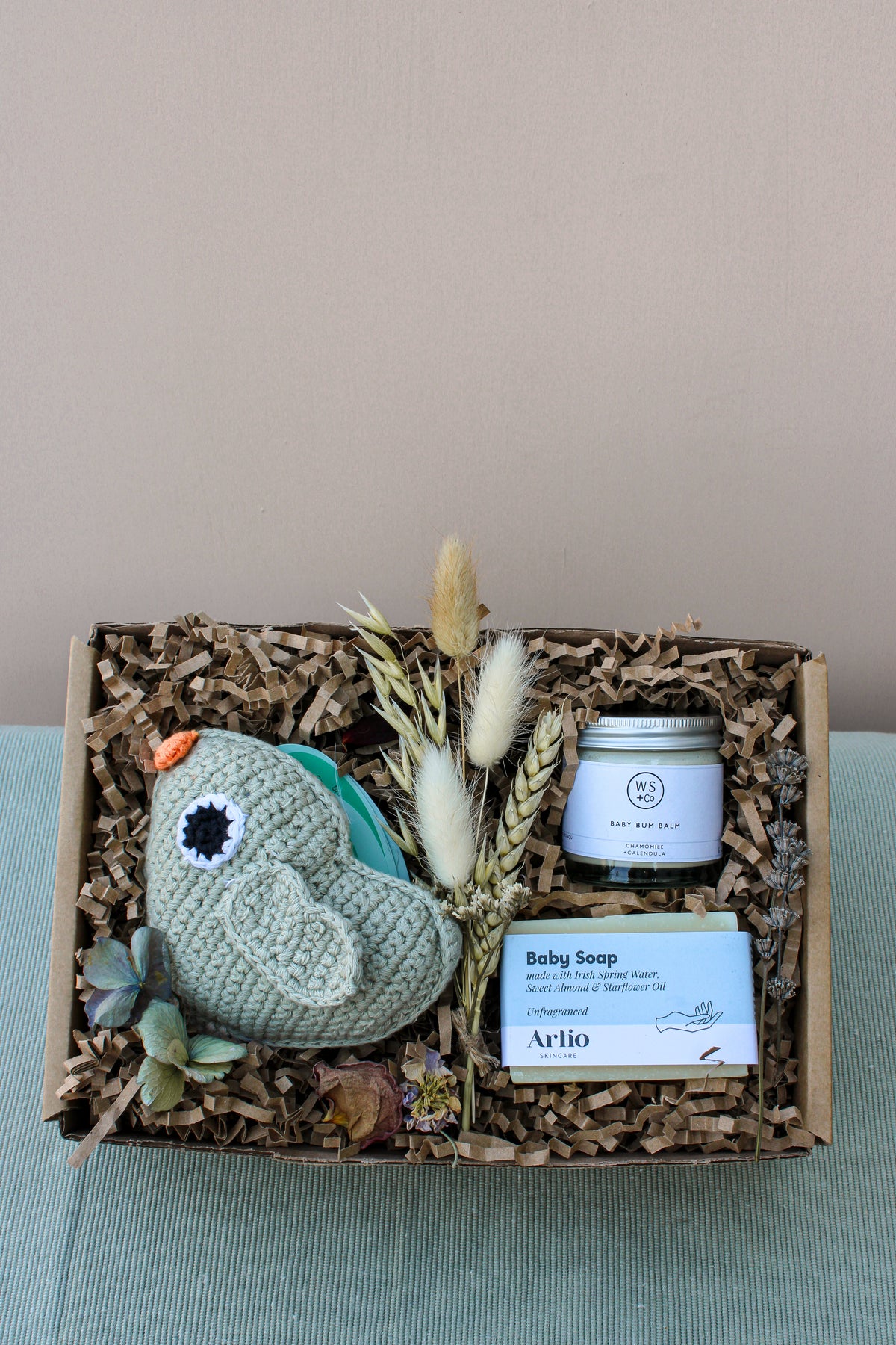The Bundle of Joy Gift Box from Wild About Flowers includes a delightful knitted grey baby shoe made from organic cotton, a small jar of calming baby balm, hypo-allergenic soap for gentle skin protection, and dried flowers, all arranged on a bed of shredded brown paper.
