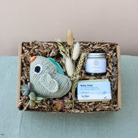The Bundle of Joy Gift Box by Wild About Flowers features a knitted bird toy, a jar of soothing baby balm, and a bar of hypo-allergenic soap, all nestled in shredded paper. The gift box is elegantly decorated with dried flowers and greenery and is displayed on a teal surface.