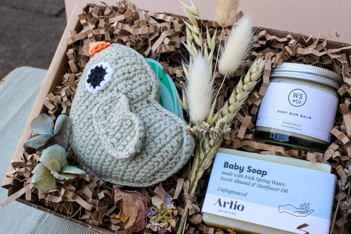 The "Bundle of Joy Gift Box" by Wild About Flowers includes a knitted bird toy crafted from organic cotton, accompanied by a small bouquet of dried flowers, a jar of soothing baby bum balm, and a bar of gentle hypo-allergenic baby soap. These delightful items are carefully arranged among shredded paper filler.