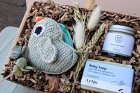 The "Bundle of Joy Gift Box" by Wild About Flowers includes a knitted bird toy crafted from organic cotton, accompanied by a small bouquet of dried flowers, a jar of soothing baby bum balm, and a bar of gentle hypo-allergenic baby soap. These delightful items are carefully arranged among shredded paper filler.