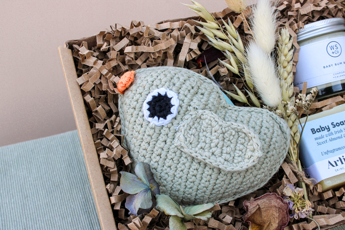 The "Bundle of Joy" gift box by Wild About Flowers includes a delightful knitted green bird toy with a black eye and an orange tuft, nestled among dried flowers and wood shavings. Accompanying the bird toy is a hypo-allergenic "Baby Soap" jar, along with other charming items discreetly tucked inside.