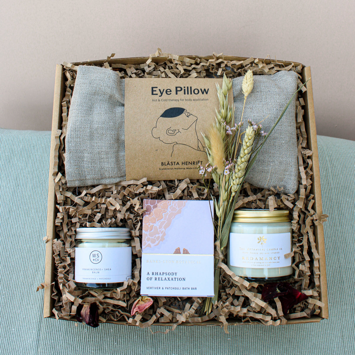 The Total Timeout Gift Box by Wild About Flowers offers relaxation with an eye pillow, a small candle, soap, and body cream. Nestled among dried flowers and crinkled paper against a light blue backdrop, it's the perfect self-care treat.