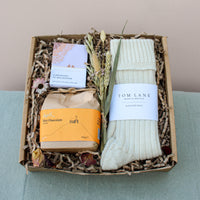 Discover Wild About Flowers' Cosy Night In Gift Box: a charming set with Swiss-style hot chocolate in yellow packaging, mint green Tom Lane alpaca bed socks, relaxation soap, and a dried floral bundle. All beautifully arranged in brown shredded paper for your perfect cozy night.