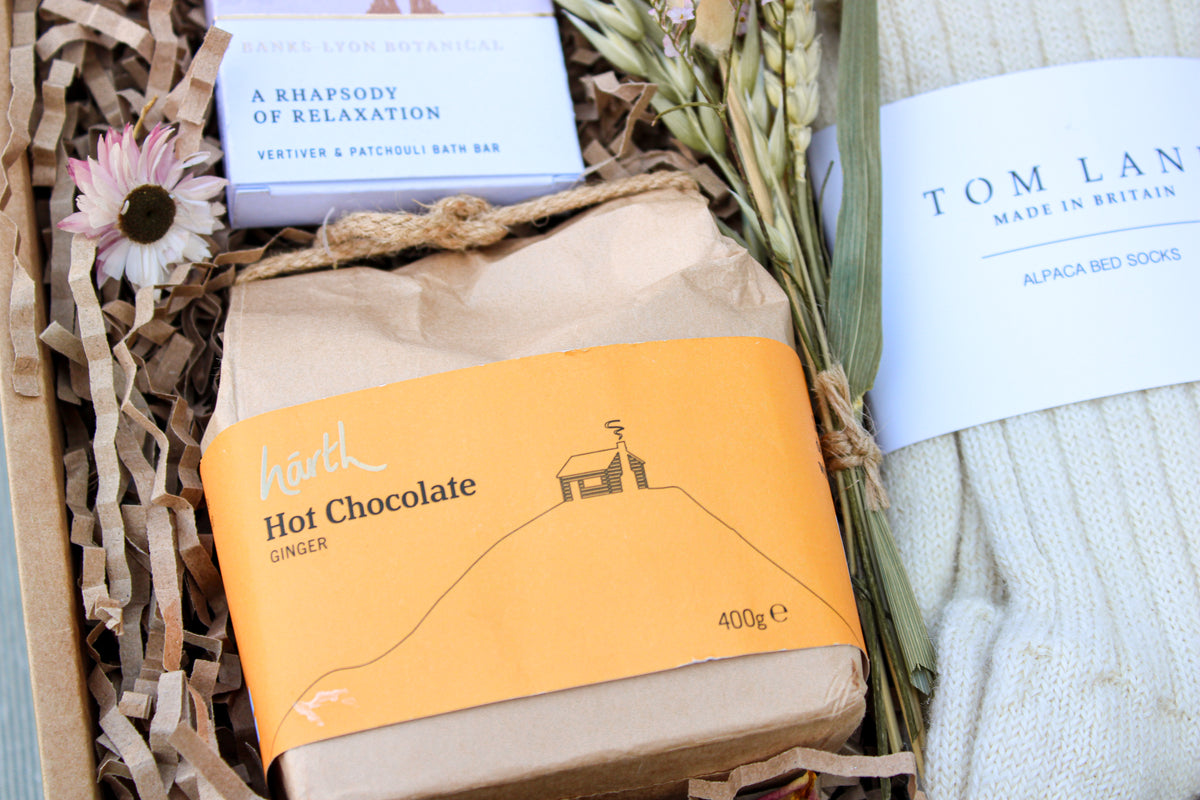 Enjoy a Wild About Flowers "Cosy Night In Gift Box" with Banks-Lyon Botanical bath soap, Harth Hot Chocolate with ginger in an orange wrapper, dried flowers, and Tom Lane Alpaca Bed Socks, all beautifully arranged on crinkled paper.