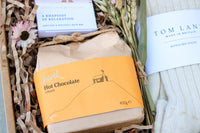 Enjoy a Wild About Flowers "Cosy Night In Gift Box" with Banks-Lyon Botanical bath soap, Harth Hot Chocolate with ginger in an orange wrapper, dried flowers, and Tom Lane Alpaca Bed Socks, all beautifully arranged on crinkled paper.