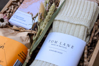 Explore the "Cosy Night In Gift Box" from Wild About Flowers, featuring cream alpaca bed socks labeled "Tom Lane Made in Britain," the book "A Rhapsody of Realisation," a yellow-brown tea packet, and elegantly framed with dried flowers and grasses for ultimate comfort.