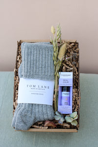 The Comfort and Calm Gift Box by Wild About Flowers features grey alpaca bed socks, a Tisserand Real Calm Roller Ball, dried flowers, and shredded brown paper for padding, all arranged on a light blue surface.