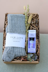 The Wild About Flowers Comfort and Calm Gift Box features cozy gray alpaca bed socks, a Tisserand Real Calm Roller Ball, dried flowers, and decorative shredded paper, beautifully arranged on a soft green surface.