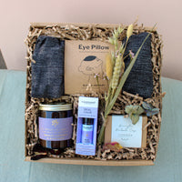 The Wild About Flowers Lavender Relax Gift Box features an eye pillow, soothing Lavender Soap Bar, French lavender & marjoram candle, Tisserand Real Calm roller ball, two dark fabric items, and dried flowers on shredded paper.