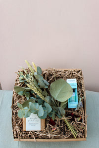 Introducing the Eucalyptus Lover Gift Box by Wild About Flowers, a relaxation gift set that includes a bundle of fresh eucalyptus leaves, soothing eucalyptus soap, and a vial of natural essential oils. These items are carefully nestled in shredded paper and beautifully displayed against a light tan backdrop on a green surface.