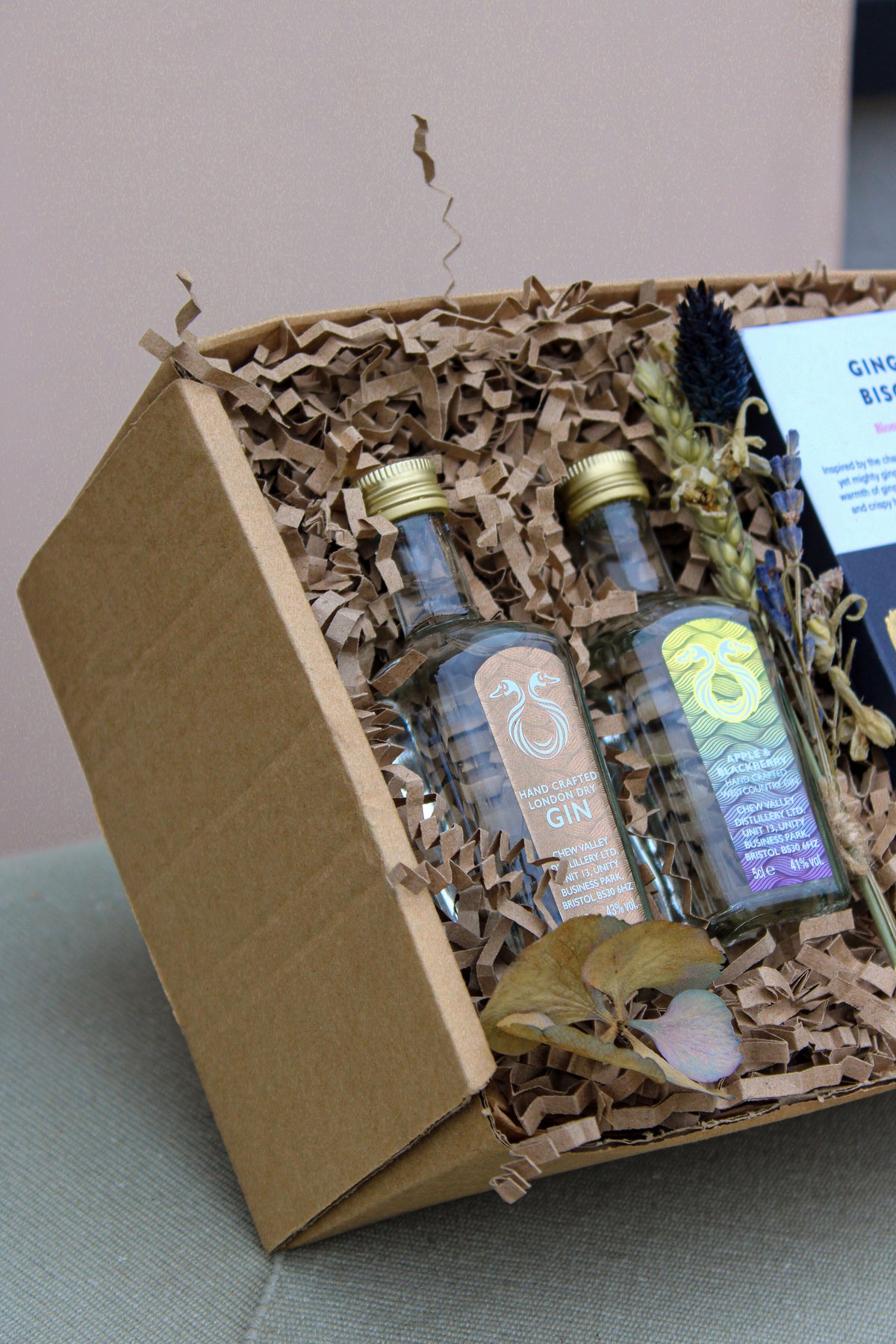 The Lovely Pair Gift Box includes two decorative gin bottles nestled in eco-friendly packaging with dried flowers adding a rustic touch, all beautifully displayed against a light pink wall.