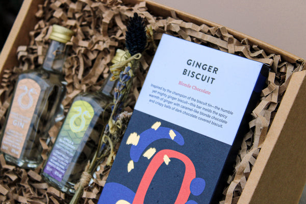 The Lovely Pair Gift Box includes two small gin bottles and a "Ginger Biscuit Blonde Chocolate" bar, elegantly packaged with eco-friendly brown crinkle paper and adorned with a charming dried flower for natural beauty.