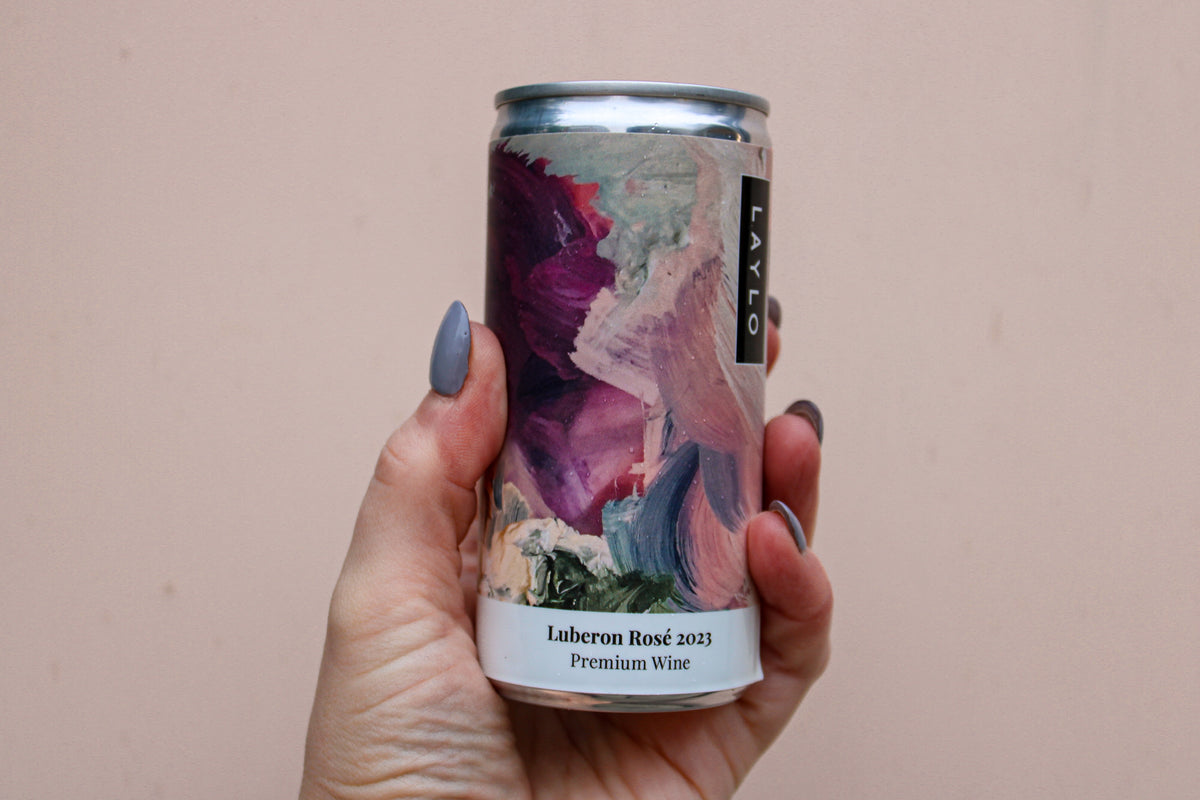 A hand holds a Luberon Rosé 2023 can from the Premium Wine Gift Set by Wild About Flowers, featuring an abstract art label in purple, pink, and beige. The person's nails are painted grayish-blue.