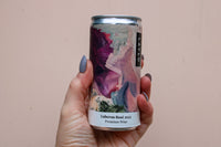 A hand holds a Luberon Rosé 2023 can from the Premium Wine Gift Set by Wild About Flowers, featuring an abstract art label in purple, pink, and beige. The person's nails are painted grayish-blue.