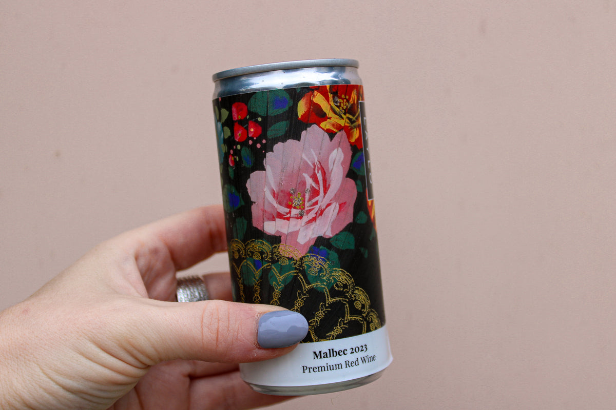 A hand with light purple nail polish holds a can of Malbec 2023 Premium Red Wine from the Wild About Flowers Premium Wine Gift Set, showcasing a floral design with a pink rose and gold accents on a dark background, reflecting its award-winning sustainability ethos.