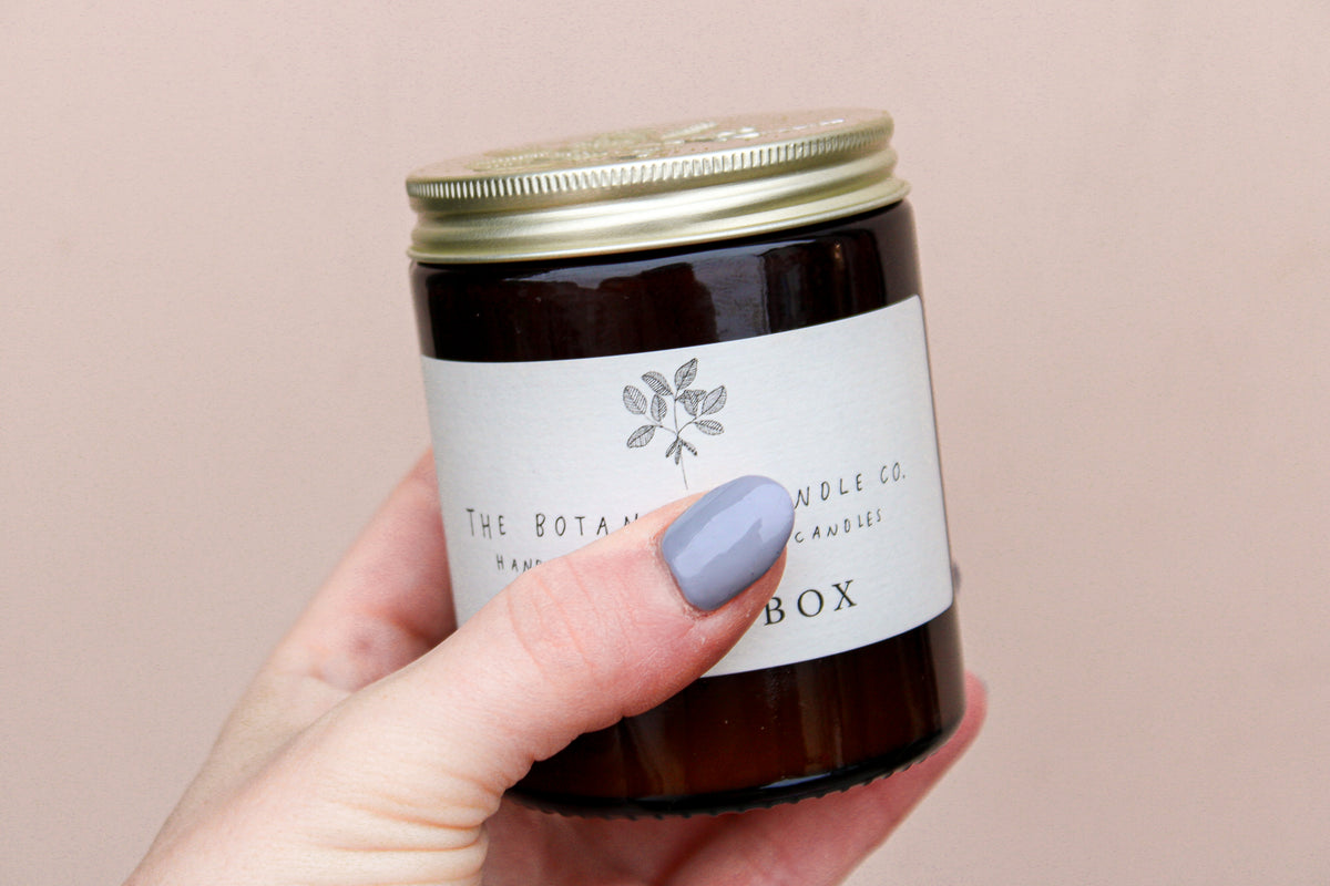 A hand with light blue nail polish holds a brown glass jar candle with a metal lid. The label has a simple leaf design and reads "Wild About Flowers." This Natural Soy Wax Candle delivers seasonal scents. The background is plain, soft beige.