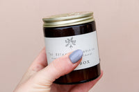 A hand with light blue nail polish holds a brown glass jar candle with a metal lid. The label has a simple leaf design and reads "Wild About Flowers." This Natural Soy Wax Candle delivers seasonal scents. The background is plain, soft beige.