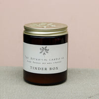 A brown glass jar candle with a gold lid sits on a light gray surface. The label reads "Wild About Flowers Natural Soy Wax Candle - Tinder Box" and features a simple plant illustration. Ethically made, this candle adds charm to any space against the soft beige background.