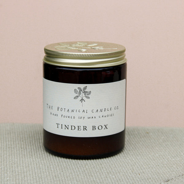A brown glass jar candle with a gold lid sits on a light gray surface. The label reads "Wild About Flowers Natural Soy Wax Candle - Tinder Box" and features a simple plant illustration. Ethically made, this candle adds charm to any space against the soft beige background.