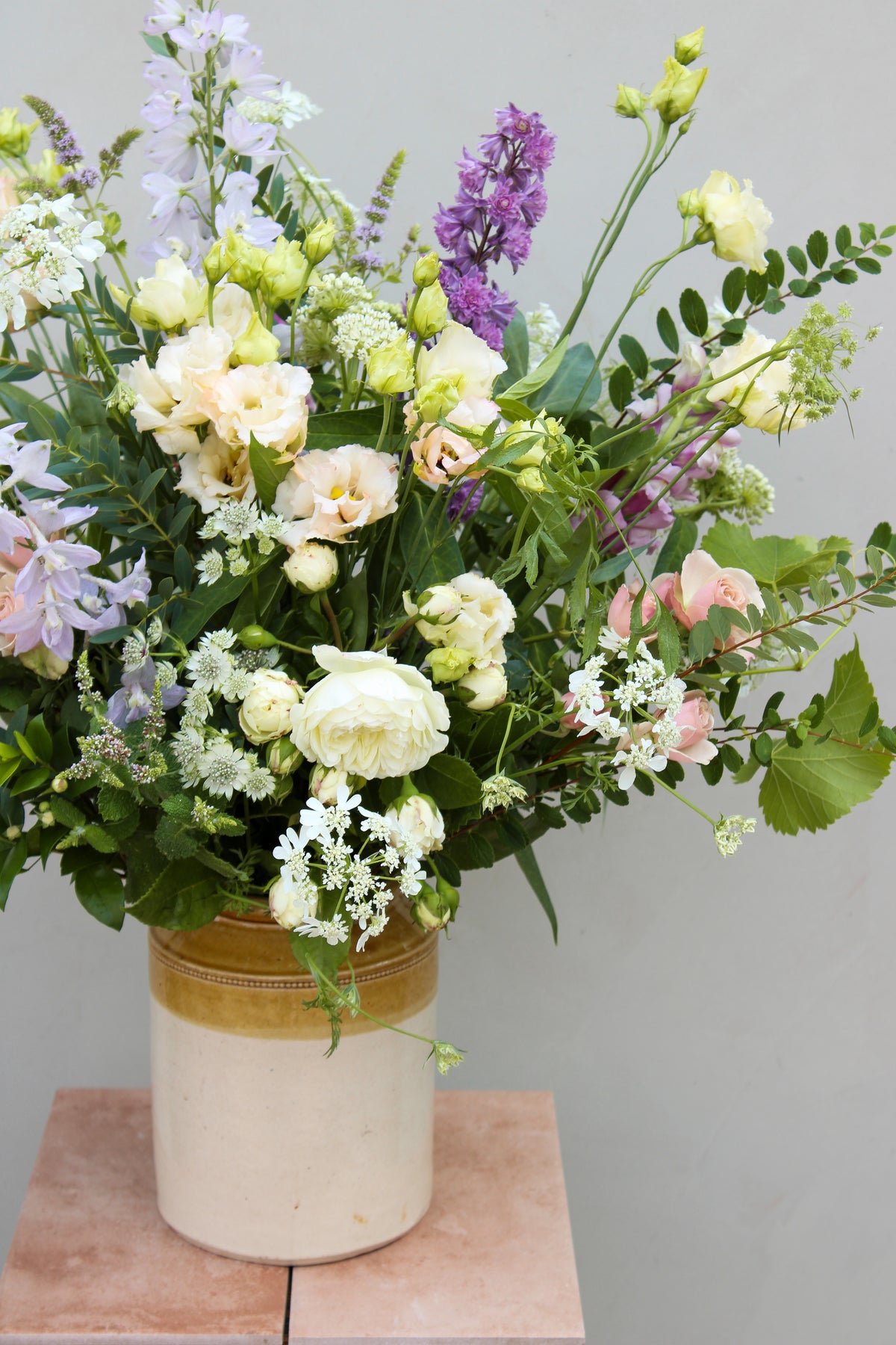A large bouquet in the Pretty pot by Wild About Flowers sits on a small table. The sustainable decor showcases white roses, lisianthus, Queen Anne's lace, and light purple blooms, complemented by lush green foliage against a simple light background.