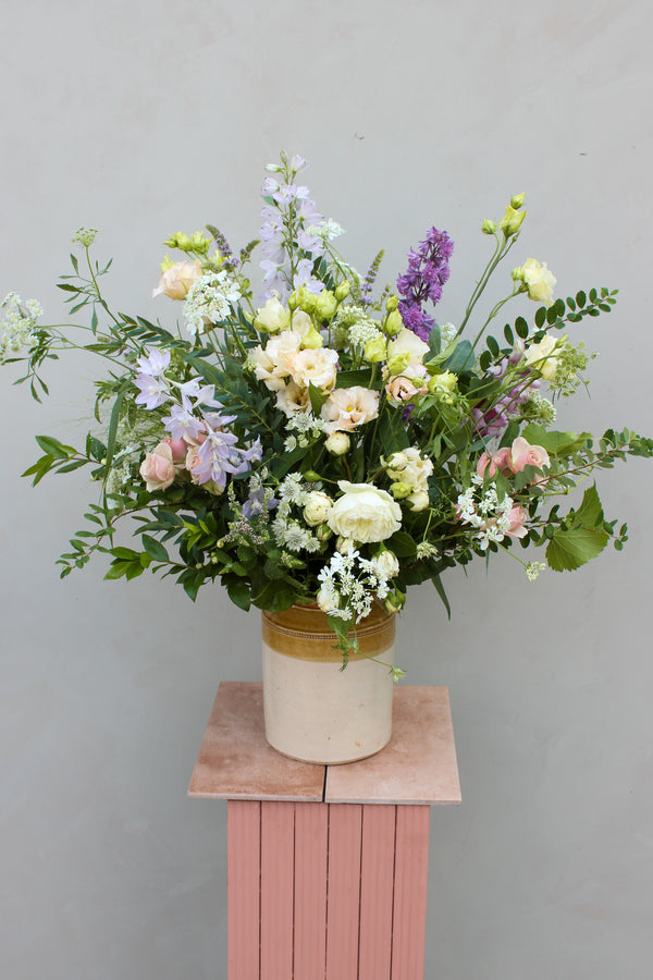 The Pretty pot by Wild About Flowers is a striking tall floral arrangement displayed in an earthenware ceramic container. It showcases a harmonious blend of white, pink, purple, and light green blossoms, enhanced by rich green foliage. The piece sits atop a pink pedestal against a light gray background, representing sustainable decor elegance.