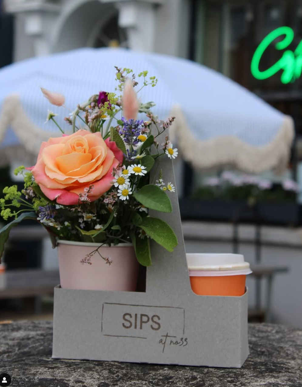 The SIPS Bundle by Wild About Flowers, featuring a cup of coffee and a small bouquet with a peach rose and daisies, is displayed outdoors with a blurred cafe backdrop and striped umbrella.
