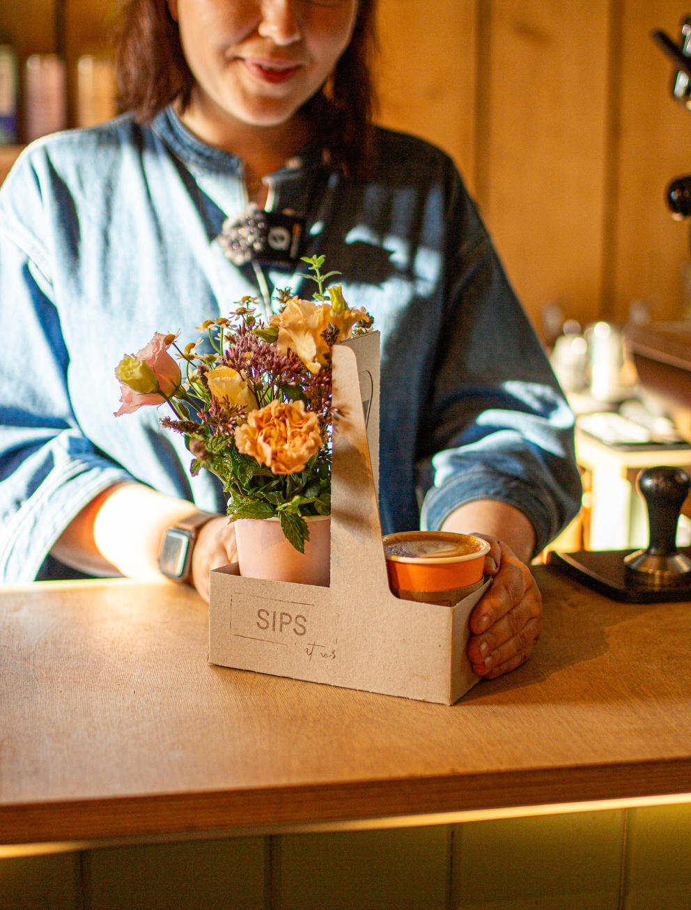 Someone in a denim shirt at a counter holds a SIPS Bundle, featuring a small bouquet and to-go coffee cup. Brand: Wild About Flowers.