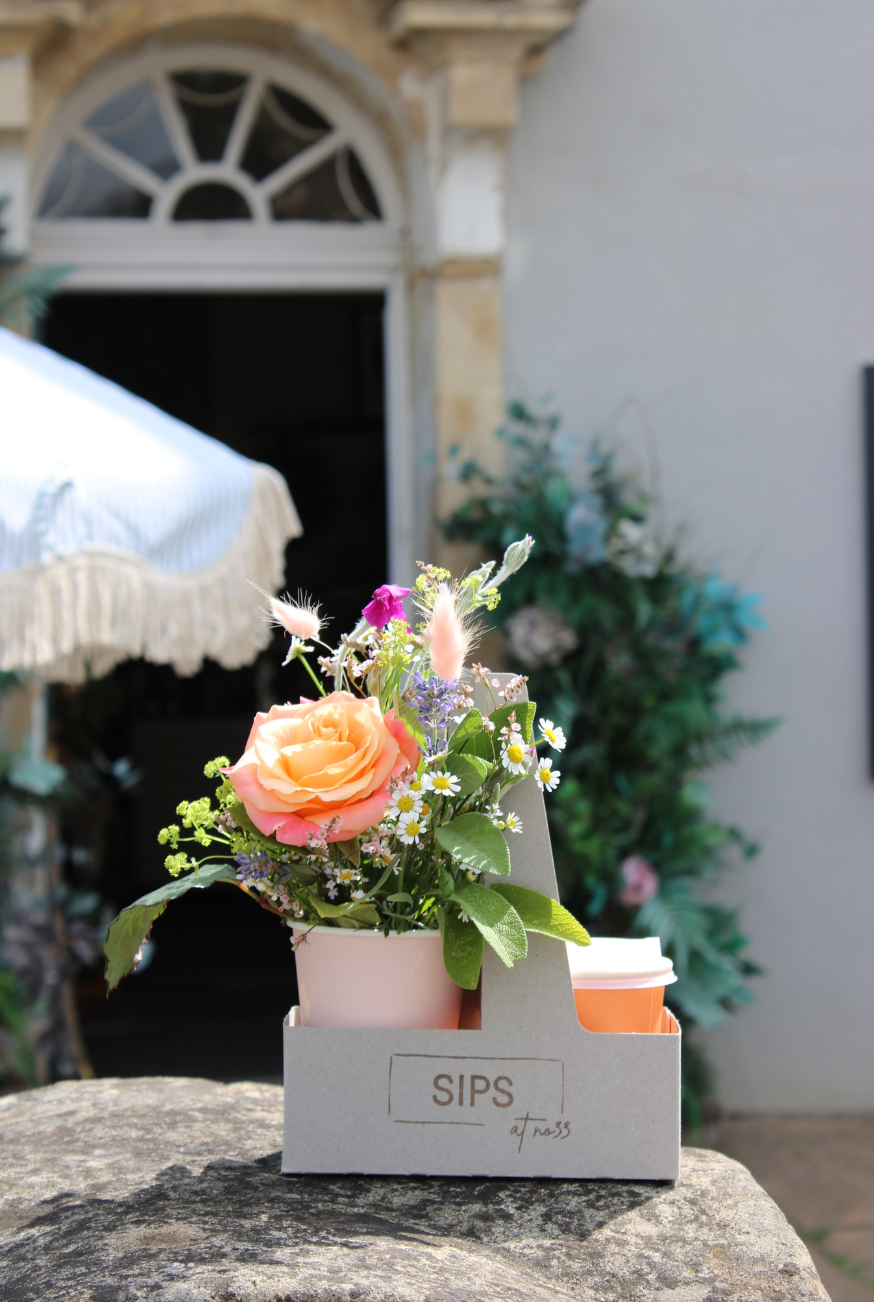 The "SIPS Bundle" by Wild About Flowers features an orange rose and wildflowers in a small cardboard holder with a disposable coffee cup, set on a stone ledge outside a building with an arched window and awning.