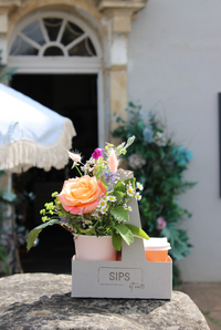 The "SIPS Bundle" by Wild About Flowers features an orange rose and wildflowers in a small cardboard holder with a disposable coffee cup, set on a stone ledge outside a building with an arched window and awning.