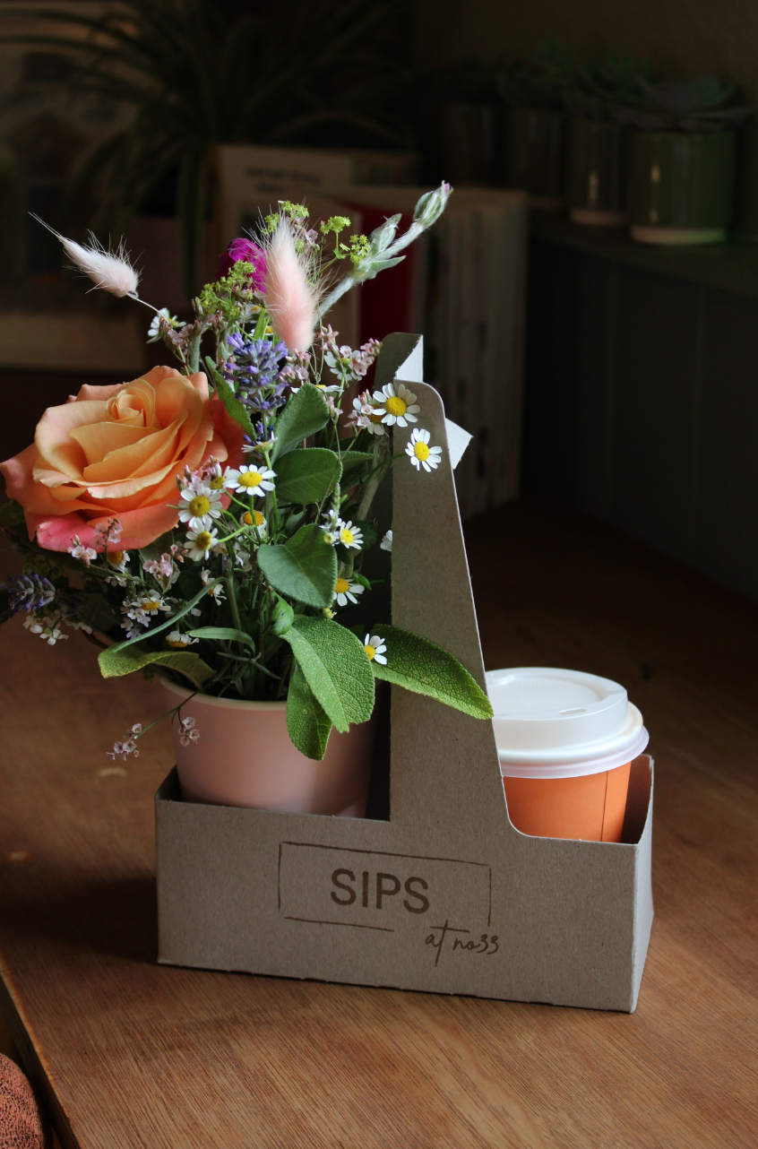 The SIPS Bundle by Wild About Flowers features a cardboard drink carrier holding a vibrant flower arrangement and an orange disposable coffee cup with a lid. Books and potted plants are visible in the background, all set on a wooden surface.