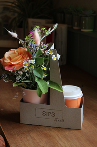 The SIPS Bundle by Wild About Flowers features a cardboard drink carrier holding a vibrant flower arrangement and an orange disposable coffee cup with a lid. Books and potted plants are visible in the background, all set on a wooden surface.